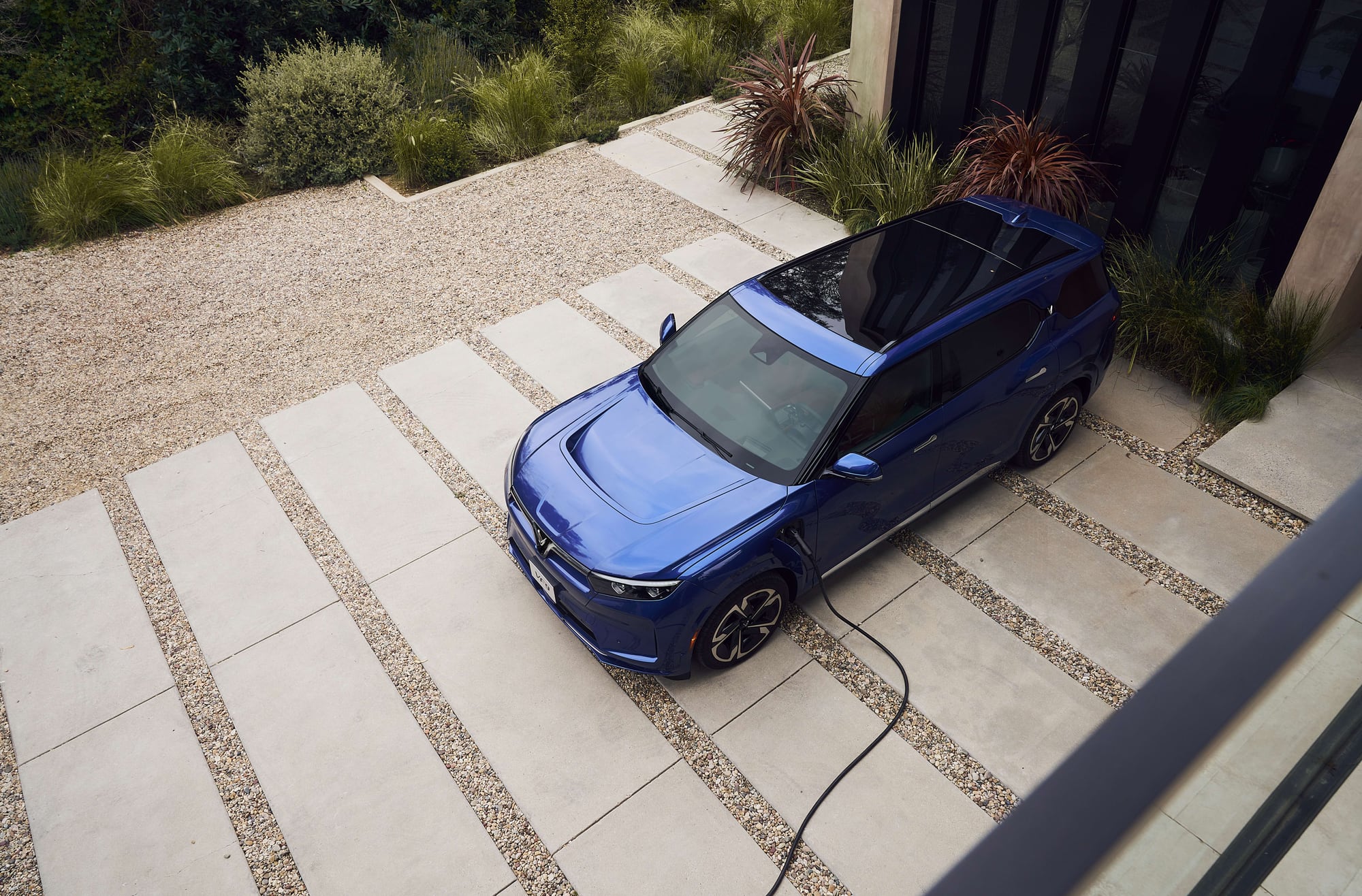 Overhead view of a blue VinFast VF 9 showing the panoramic glass roof and roof rails, charging cable connected